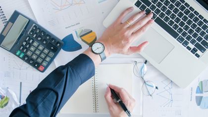 Hand on a desk with a laptop, calculator and documents